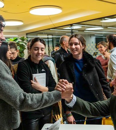 Two men shaking hands in a bustling office environment, surrounded by bystanders.