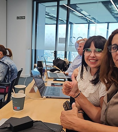 Two women smiling and posing for a photo in an office setting with computers and other people in the background.