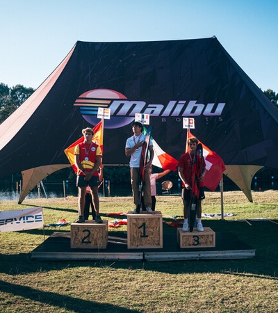 Three athletes stand on a podium at an outdoor sporting event under a large tent with the Malibu logo.