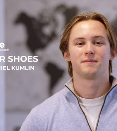 A young man smiling in front of a world map with the text 'IN THEIR SHOES with Daniel Kumlin' at IE University.