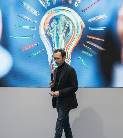 A person is standing in front of a large screen displaying a lightbulb surrounded by various creative tools.