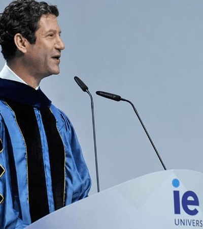 A man in academic regalia delivering a speech at a podium with 'IE University' visible.