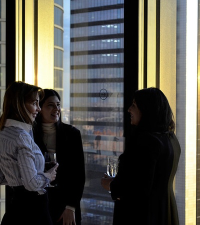 Three women conversing in an office setting with city skyline in the background at dusk