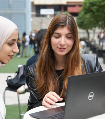 Two women are collaborating over a laptop in an outdoor setting.
