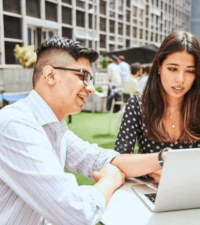 Two professionals working on a laptop outdoors in a casual office setting.