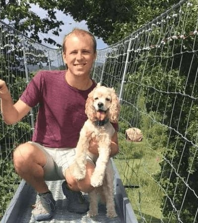 A man and a dog sitting together on a suspension bridge outdoors, both smiling towards the camera.