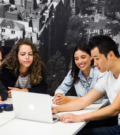 A group of four young adults is engaged in a collaborative discussion around a laptop in a modern office setting.