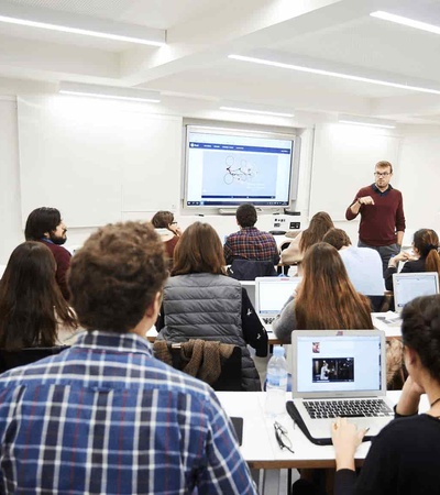 A lecturer explaining to students in a classroom equipped with laptops and a projector screen.