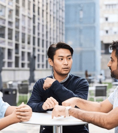 Three people having a conversation at a table outdoors, with modern buildings in the background.