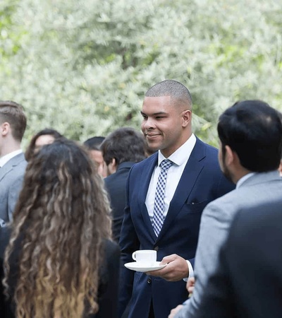 A man in a suit with a tie is smiling while holding a coffee cup, interacting with other people at an outdoor gathering.