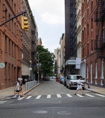 A typical urban street scene with people walking across the crosswalk, surrounded by historic brownstone buildings and parked cars under a cloudy sky.