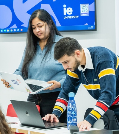 A student assists another student with a laptop in a university classroom.