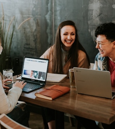 Three friends are laughing together while working on laptops at a wooden table.