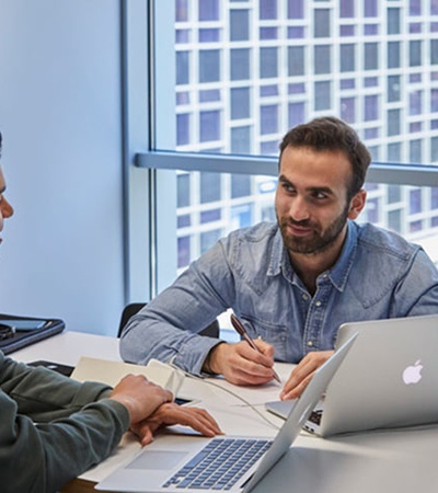 Two men are engaged in a discussion over a laptop at a table.