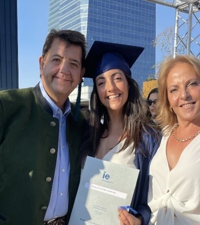 A family celebrates a graduation ceremony outdoors, with a young woman in a blue cap and gown holding a diploma, flanked by an older man and woman.