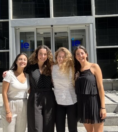 Four young women smiling in front of a building with large glass doors and the 'IE' logo above them.