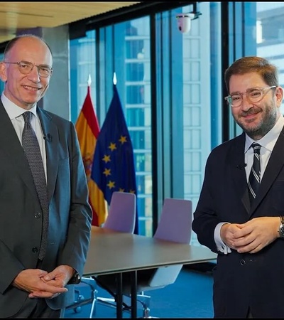 Two men in suits standing in an office with a modern design aesthetic, including a visible background featuring flag displays and multimedia screens.