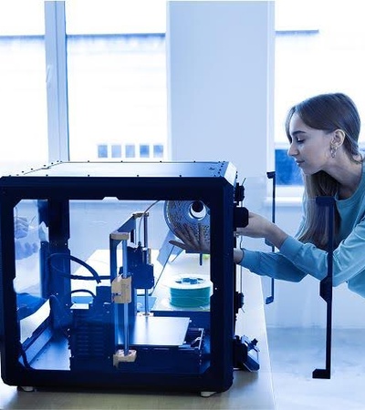 A woman operates a 3D printer while a man works at a nearby table in a bright room.