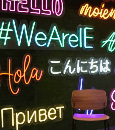 A wall decorated with various greetings in multiple languages in neon lights, accompanied by a single stool in front.