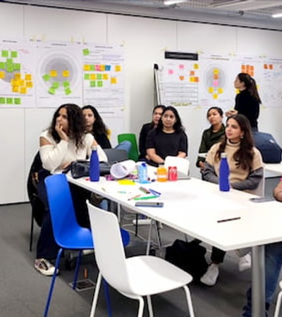 A woman presenting to a group of seated people in a colorful workshop room with posters on the walls.