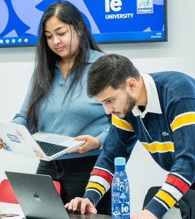 Two students are engaged in a discussion while working on their laptops in a classroom.