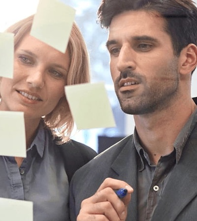 A man and a woman in a modern office setting, brainstorming using sticky notes on a glass wall.