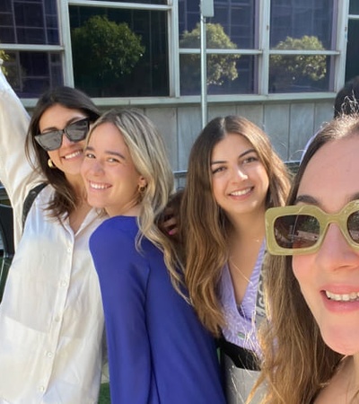 Four women smiling for a selfie outdoors, one of them raising her hand in excitement.