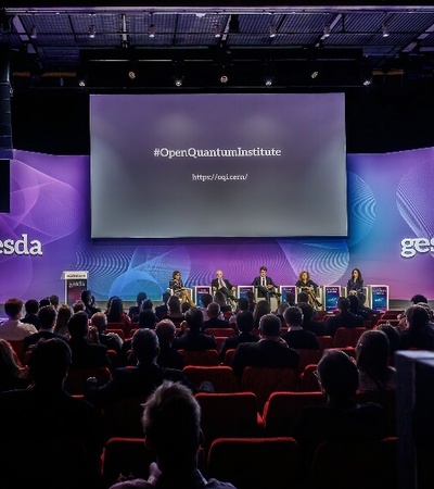 A panel discussion at a conference with a crowd watching, featuring a large screen displaying the hashtag '#OpenQuantumInstitute' and other visual graphics.