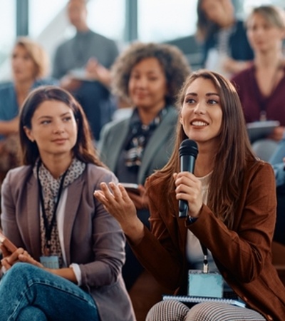 A woman in a brown jacket is speaking into a microphone during a seminar while sitting among an attentive audience.