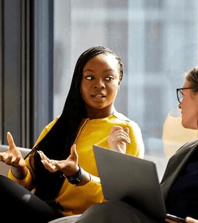 Two professional women engaged in a conversation in an office setting, one of them gesturing while holding a laptop.