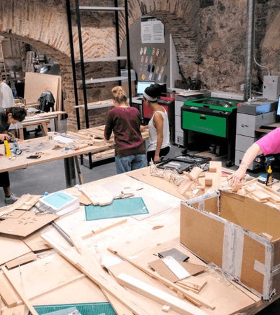 People are working collaboratively in a spacious workshop with various tools and materials on their desks.