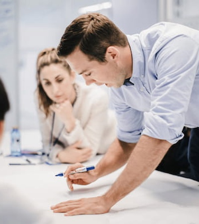 A man in a blue shirt is leaning over a table, explaining something to two colleagues in an office setting.
