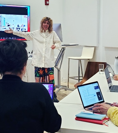 A woman is giving a presentation in a modern classroom setting, pointing towards a screen displaying video calls with other participants.