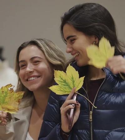 Two young women smiling and holding autumn leaves in a room with mannequins.