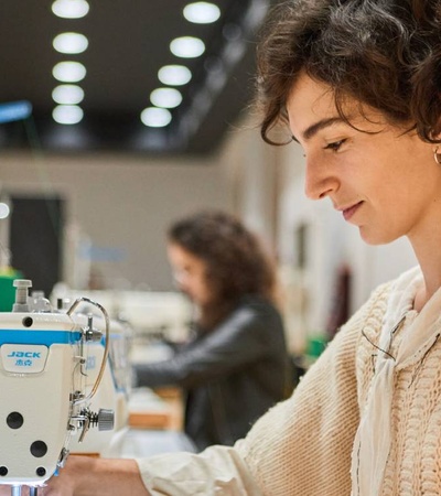 A woman operating a sewing machine in a workshop setting with other people in the background.