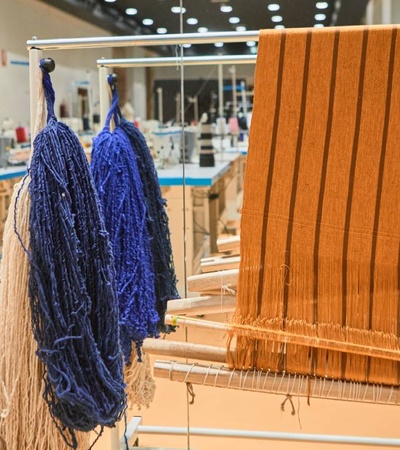 A textile workshop with a weaving loom and yarns of various colors in the foreground, and workers in the background.