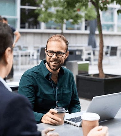 Three people are engaged in a discussion at an outdoor table, one of them is using a laptop.