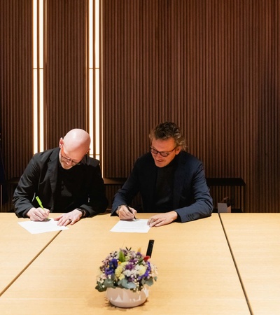 Two men signing documents at a table in a modern office with warm lighting and flags in the background.