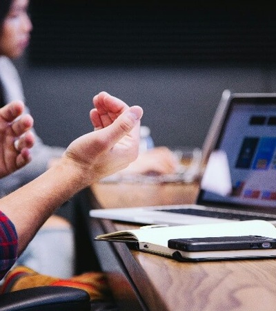 A person in a plaid shirt gestures during a meeting with a laptop and smartphone visible on the table.