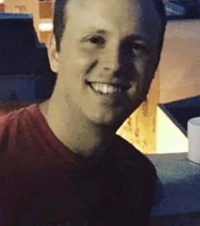 A smiling man in a red shirt sitting at a table with a white cup visible in the background.