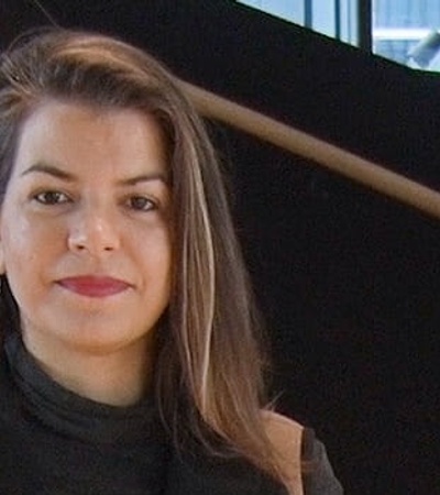 A woman standing on a staircase with a subtle smile, wearing a black turtleneck.