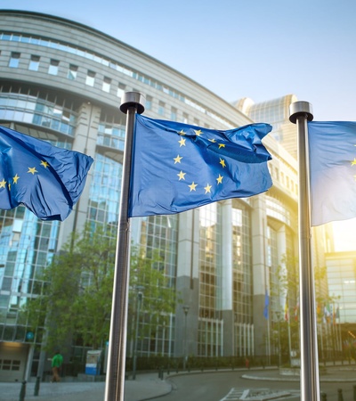 Three European Union flags flutter in front of a modern glass building on a sunny day.