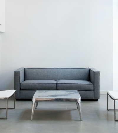 A minimalistic living room with a gray sofa, two white chairs, and a marble table against a backdrop of white walls and large windows.