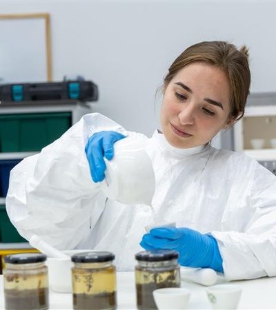 A scientist in a lab coat is carefully pouring a substance into a container while examining samples.