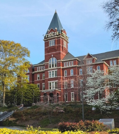 A historic brick building with a tower, surrounded by trees and flowering plants in a park-like setting.