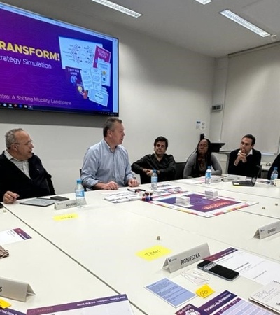 A group of business people engaged in a meeting around a table with documents and a digital presentation in the background.