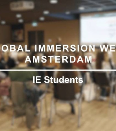 A blurry image of people sitting in a conference room with text indicating 'Global Immersion Week Amsterdam' and 'IE Students'.