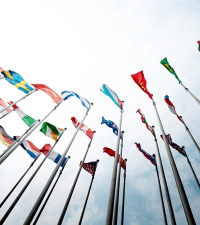 A collection of various national flags fluttering on poles against a blue sky with white clouds.