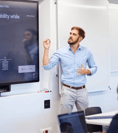 A man with a beard and blue shirt is giving a presentation in a classroom with a digital screen displaying text.