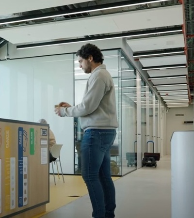 A man stands in an office hallway next to recycling bins, using his smartphone.